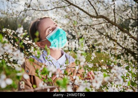 Woman allergic suffering from seasonal allergy at spring, posing in blossoming garden at springtime, wearing medical mask among blooming trees. Spring allergy concept Stock Photo