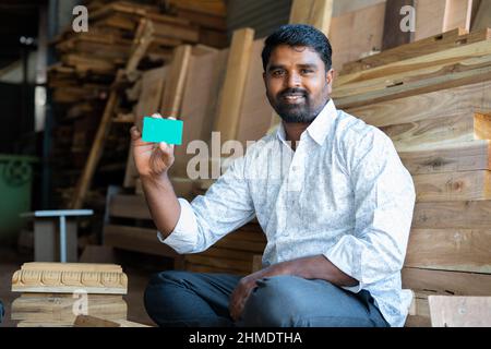 Smiling indian carpenter showing empty green card mockup by looking at camera - concept of employee id card, advertising, skilled labour and Stock Photo