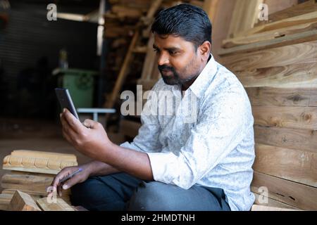 smiling carpenter busy using mobile phone at carpentry shop - concept of technology, social media, surfing internet and checking deisgns Stock Photo