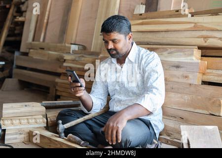 young indian carpainter or craftperson busy using mobile phone at workplace - concept of technology, seeing or checking design and skilled labour. Stock Photo