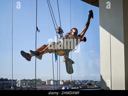 Industrial mountaineering worker hanging on climbing rope and cleaning window of high-rise building. Man using safety lifting equipment while wiping skyscraper window with rag. Stock Photo