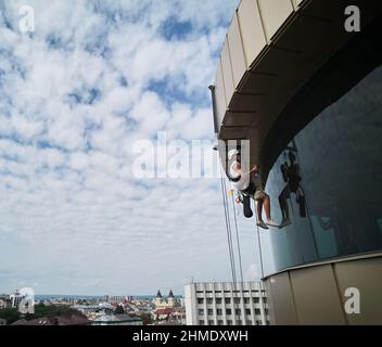 Industrial mountaineering worker hanging on climbing rope and cleaning glass window of skyscraper. Male cleaner using safety lifting equipment while washing window of high-rise building. Stock Photo