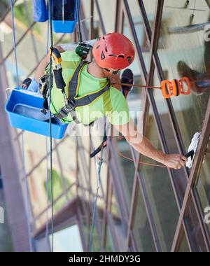 Industrial mountaineering worker washing glass windows of high-rise building, hanging on safety climbing rope. Man window cleaner in protective helmet cleaning skyscraper facade. Top view. Stock Photo