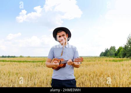 Singer. Young beautiful caucasian man playing ukulele outside. Handsome hipster guy plays ukulele guitar on nature background. Male in a cowboy hat sm Stock Photo