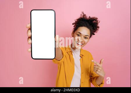 Smartphone mock-up. Excited happy african american girl, holding cellphone in hand with blank white screen for presentation, pointing finger at it, stand over isolated pink background,smiling joyfully Stock Photo