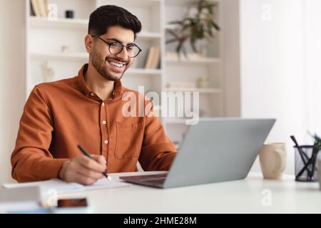 Smiling Arab man in glasses using laptop and writing Stock Photo