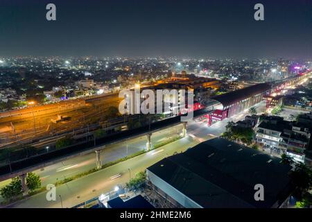 aerial drone shot showing elevated metro train station and tracks over busy street with light trails from traffic and cityscape in gurgaon, jaipur Stock Photo