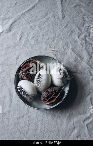 Moody spring still life scene. Easter composition. Bowl of folded paper ornaments and hen eggs decorated by feather of guinea fowl bird. Gray linen Stock Photo