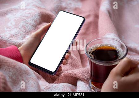 Woman sitting on bed and using mobile phone with blank white desktop screen. Mockup image of hand holding smartphone. Stock Photo