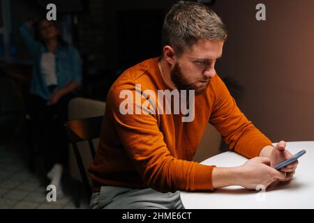 Busy addicted young man using mobile phone sitting at table in dark living room on background of girlfriend sitting on chair. Stock Photo