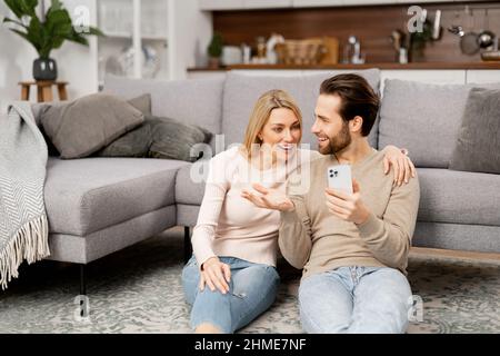 Excited and overjoyed caucasian young couple sitting near the couch at home and holding smartphone. Man and woman cant believe at something, celebrating good news or win, received money on account Stock Photo