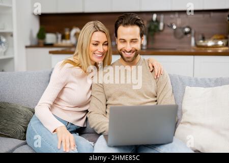 Blonde woman and man looking at the laptop screen while sitting on the sofa and watching comedy movies. Overjoyed couple in love spending leisure time online with laptop at home Stock Photo