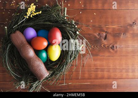 Easter eggs and chocolate bunny in a nest of branches on dark wooden background. Copy space. Top view. Stock Photo