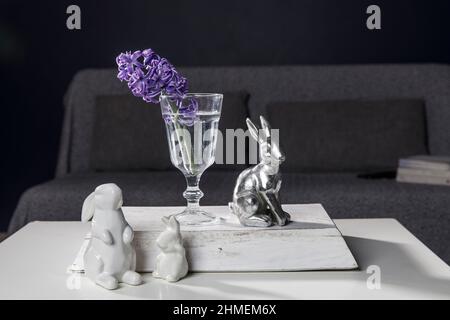 Ceramic figurines of Easter bunnies of different sizes on the table. Blue and white hyacinths in glass cups on a dark background. Easter design Stock Photo