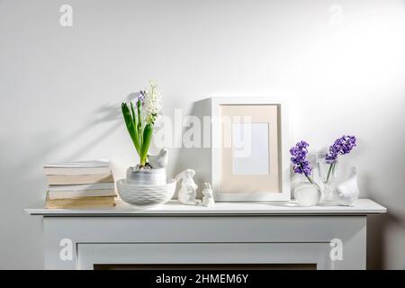 White hyacinth in a large porcelain bowl, books, figurines of hares and a bird, an empty photo frame are on the fireplace against the white wall. Layo Stock Photo