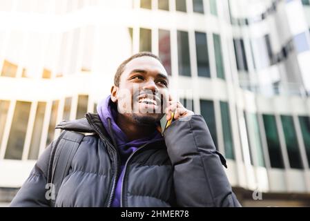 From below African American man in outerwear smiling and looking away. while talking on mobile phone on blurred background of modern building on city street Stock Photo