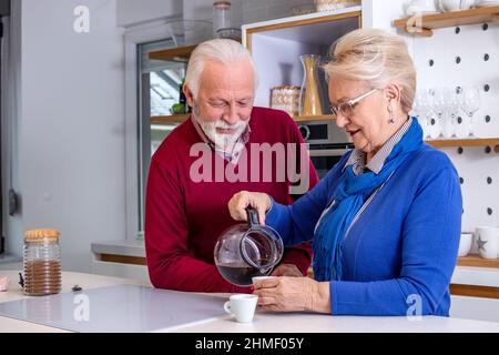 Happy senior couple preparing their morning coffee in the kitchen, smiling and talking to each other. Stock Photo