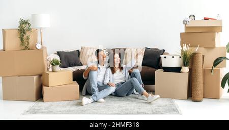 Happy spouses planning interior design of new home. Excited joyful mixed race couple in love, sitting on the floor in the living room in their new home, cardboard boxes with things are standing nearby Stock Photo