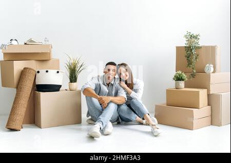 Happy in love couple, caucasian woman and hispanic man, stylishly dressed, sit in their first own house on the floor, there are card boxes with things around, they smiling, invested in real estate Stock Photo