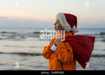 Portrait of a cute boy in a cap of St. Nicholas, with a bag of Christmas and New Year's gifts on the background of the sea, the guy holds a bag of gif Stock Photo