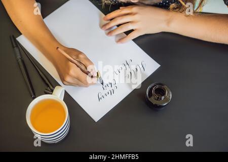 Calligrapher Young Woman writes phrase on white paper. Inscribing ornamental decorated letters. Calligraphy, graphic design, lettering, handwriting Stock Photo