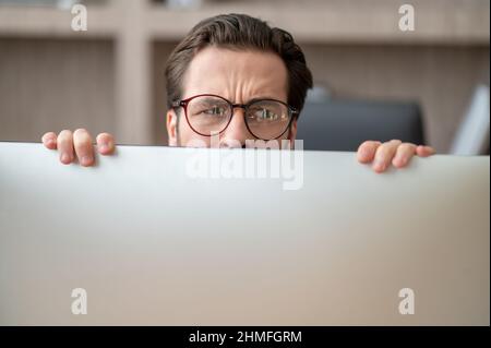 A dark-haired man hiding behind the computer and looking stressed Stock Photo