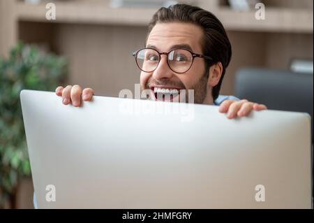 A dark-haired man hiding behind the computer and smiling Stock Photo