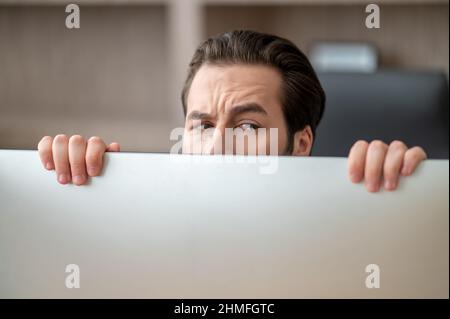 A dark-haired man hiding behind the computer and looking stressed Stock Photo