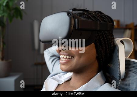Happy relaxed afro woman in VR googles using virtual reality to relax and reduce stress at workplace Stock Photo