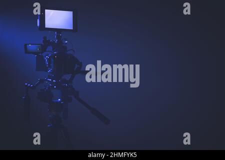 Modern Digital Cinema Camera with External Display Staying on a Tripod Inside Dark Blue Illuminated Studio. Right Side Copy Space Stock Photo