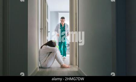 Worried doctor running to help mental disorder female patient sitting on floor in hospital corridor Stock Photo