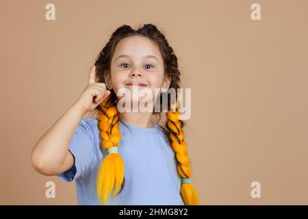 Little smart girl with kanekalon braids of yellow color showing finger having aha moment because of great idea appearing in head on beige background Stock Photo