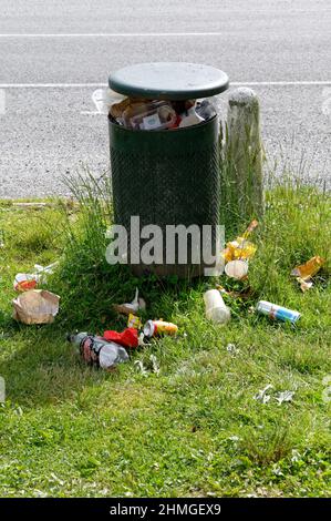 People have discarded rubbish despite the rubbish bin being full. The litter is on the ground surrounding the bin. Stock Photo