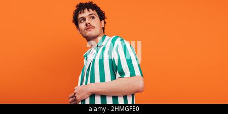 Young queer man looking away thoughtfully in a studio. Confident young generation z hipster standing alone against an orange background. Stock Photo
