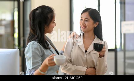 Two female colleagues are having a chill talk while sipping coffee on their coffee break in the office's snack room. Friendly businesswomen concept. Stock Photo