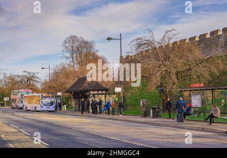 A bus stop and buses underneath historic city walls. Passengers wait while others alight a bus and a blue sky with clouds is above. Stock Photo