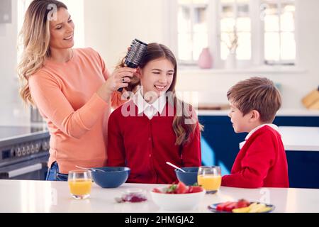 Mother Brushes Daughter's Hair As Children In School Uniform Eat Breakfast At Home Stock Photo