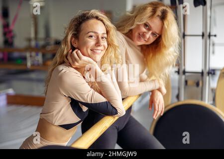 Portrait of smiling young woman looking at camera while standing near ballet barre after pilates workout together with her instructor in stretching Stock Photo