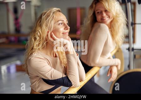 Portrait of cute young woman, dancer smiling away while standing near ballet barre together with her friend in stretching studio Stock Photo