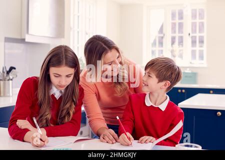 Mother Helping Children In School Uniform Doing Homework At Kitchen Counter Stock Photo