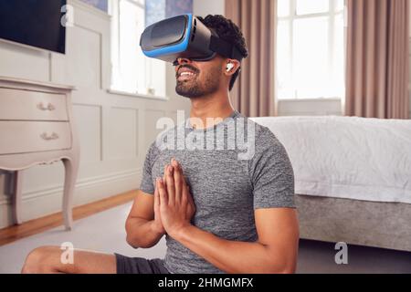 Man Wearing Virtual Reality Headset And Earphones Sitting On Yoga Mat In Bedroom At Home Stock Photo