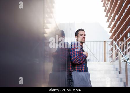 Confident handsome bearded male in jeans jacket leaning on outdoor wall Stock Photo