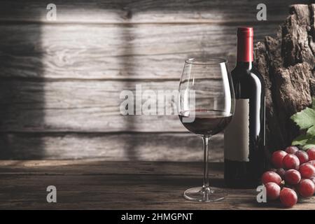 Elegant glass of wine and red wine bottle with grapes and shadows next to the window  on dark wood background with copyspace. Stock Photo