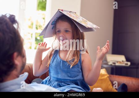 Father And Daughter Lying On Sofa At Home As Girl Puts Reading Book On Head Stock Photo