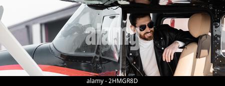 bearded pilot in stylish sunglasses and leather jacket smiling while sitting in cockpit of helicopter, banner Stock Photo
