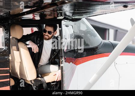 bearded pilot in stylish sunglasses and leather jacket smiling while sitting in cockpit of helicopter Stock Photo