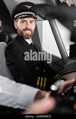 bearded pilot in uniform and cap smiling near blurred co-pilot Stock Photo