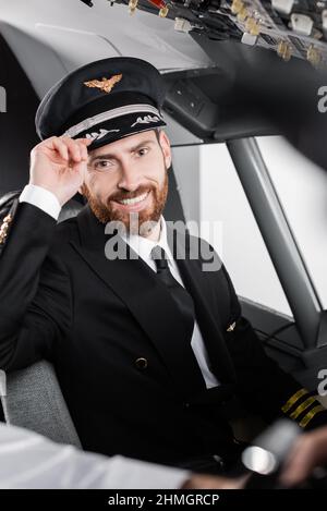 bearded pilot in uniform adjusting cap and smiling near blurred co-pilot Stock Photo