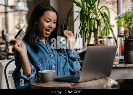 Black excited woman using laptop and holding credit card while sitting in cafe Stock Photo