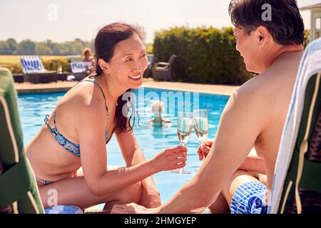 Mature Couple On Loungers Relaxing By Swimming Pool On Summer Vacation Drinking Champagne Stock Photo